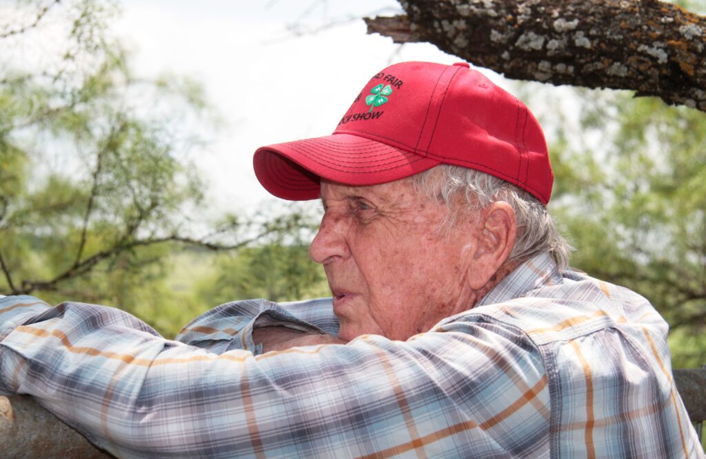 A man in plaid shirt and red hat leaning on tree.