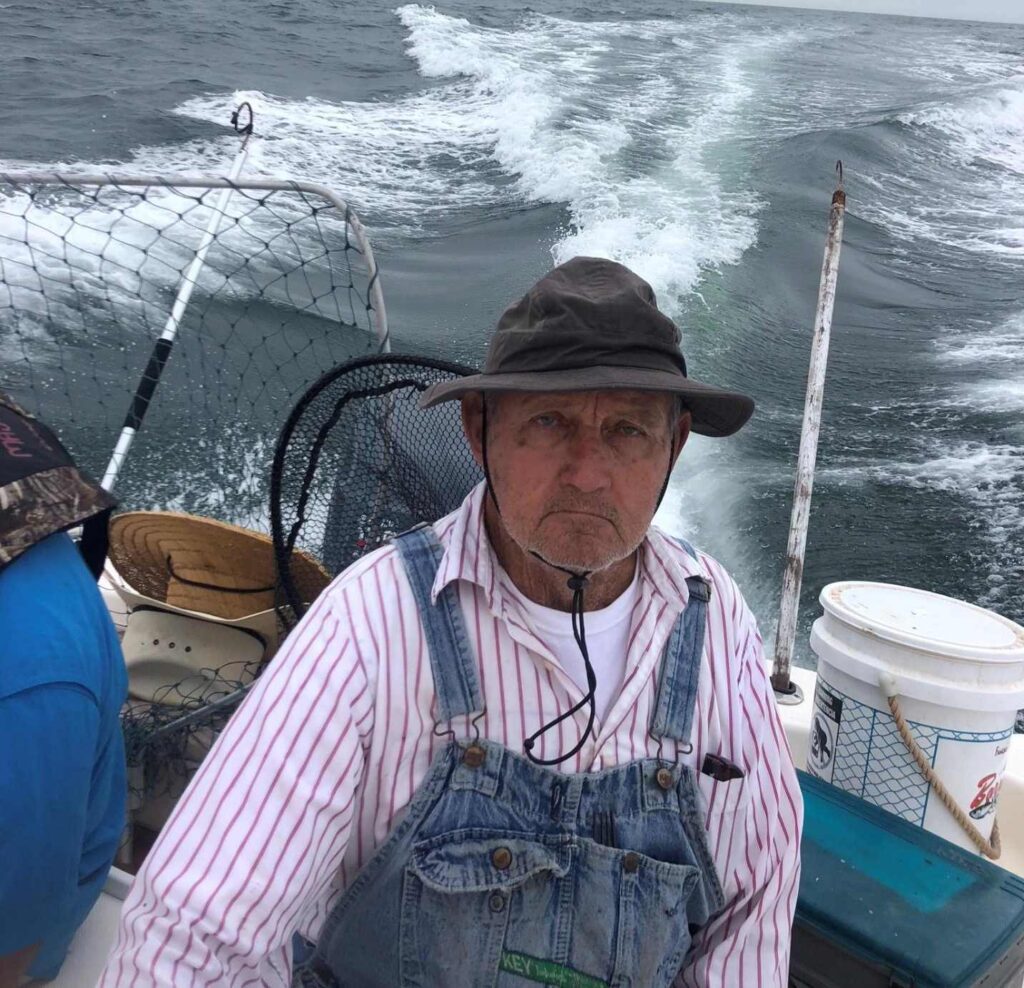 A man in an overalls and hat standing on the deck of a boat.