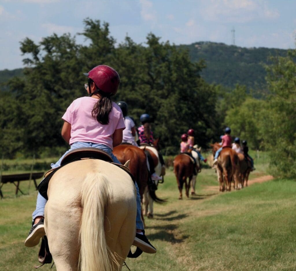 Children riding horses on a trail through a scenic landscape.