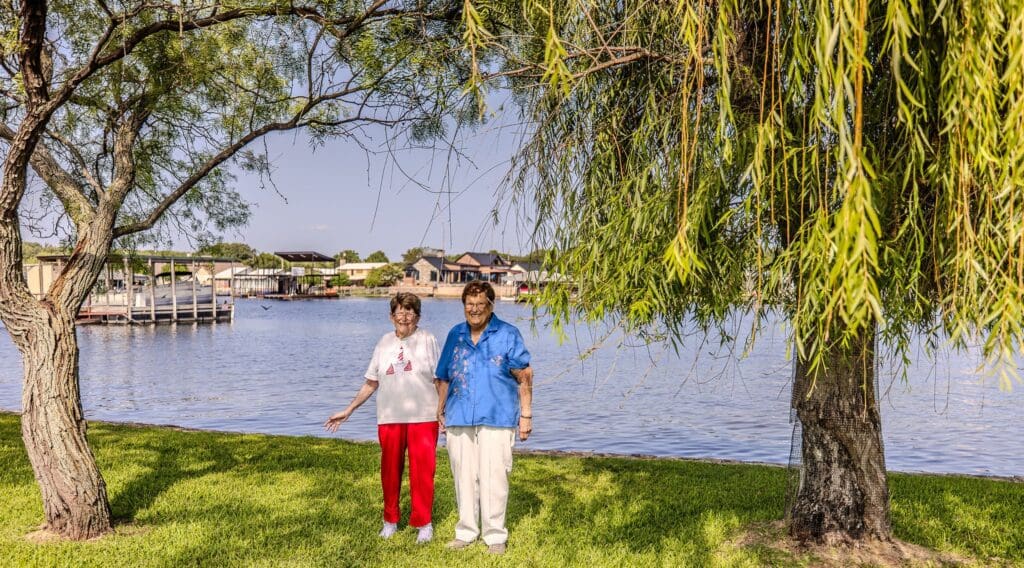 Two people standing by a lake under a willow tree on a sunny day.