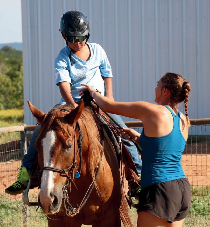 A woman helps another woman ride a horse outdoors.