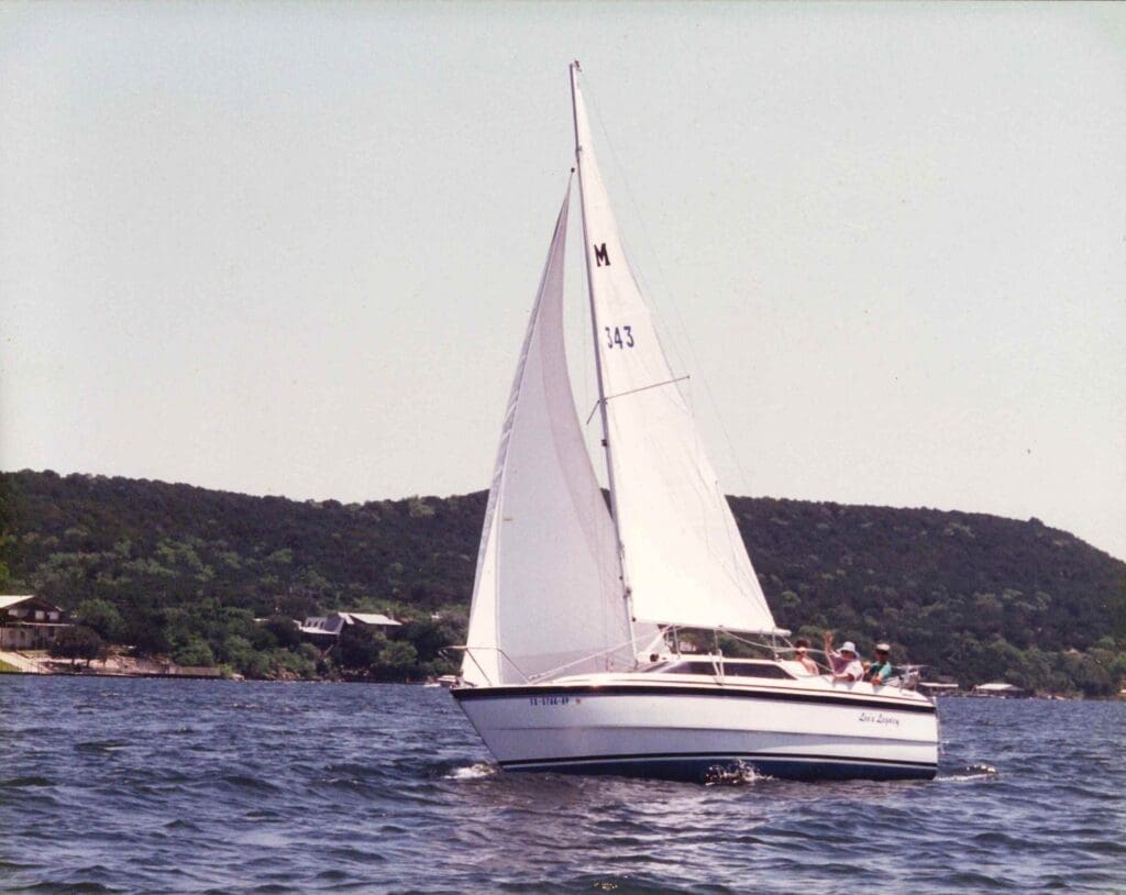 Sailboat with white sails gliding on calm water near a green shoreline.