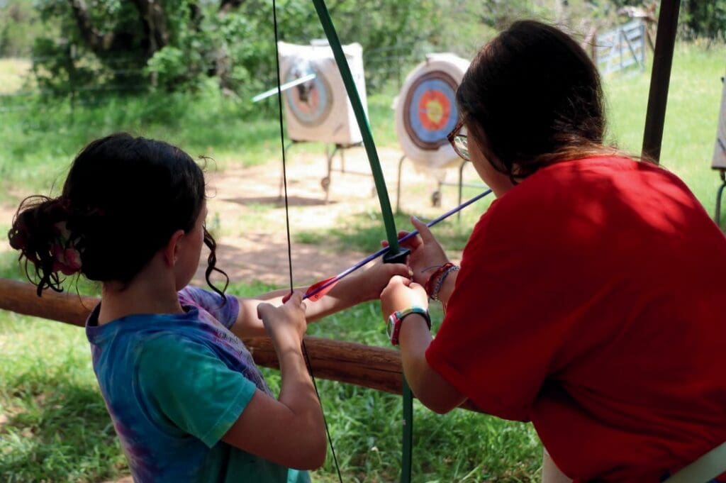 An adult teaches a child archery outdoors.