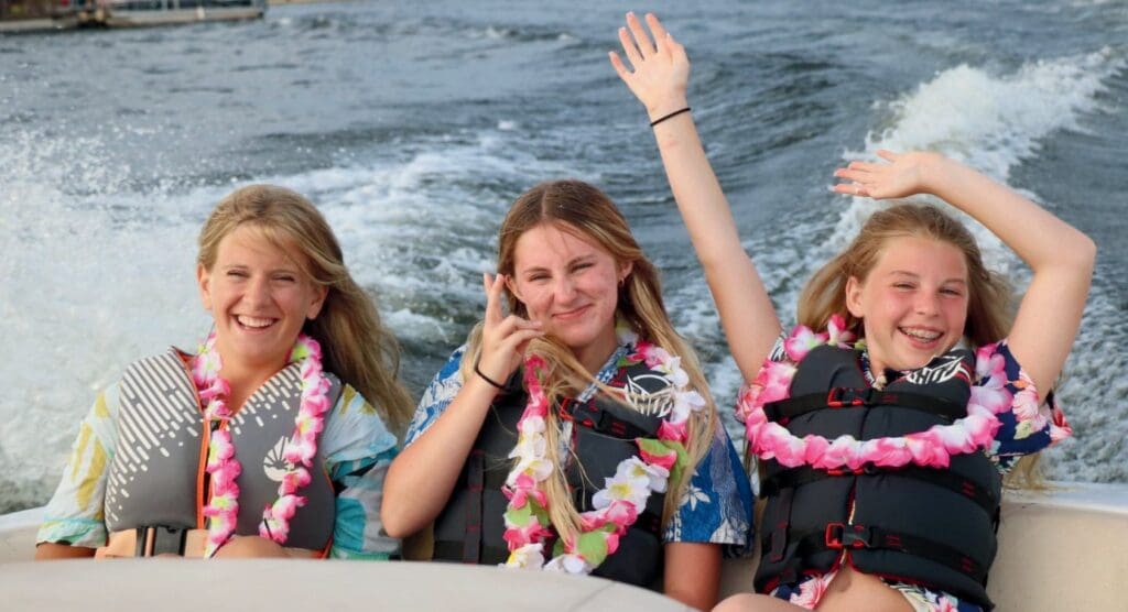 Three girls wearing leis and smiling on a boat with ocean waves behind.